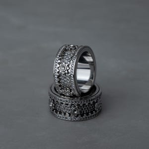 May include: A silver-toned metal ring featuring a wide band design with a central row of gear-shaped motifs. The surface is encrusted with numerous small, clear, sparkling stones, and the gear centers are accented with single, larger, clear stones.