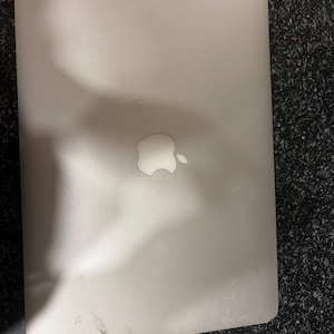 May include: A silver laptop with a smooth, metallic finish and a rounded rectangular shape. The center of the lid features a prominent, reflective Apple logo. The surface of the device shows minor signs of use with light smudges and surface marks.