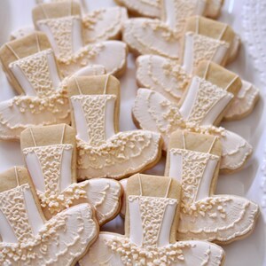 May include: A collection of ballet tutu cookies, decorated with white icing. The cookies are shaped like tutus, with detailed bodice and skirt designs. The cookies are arranged on a white, decorative plate.