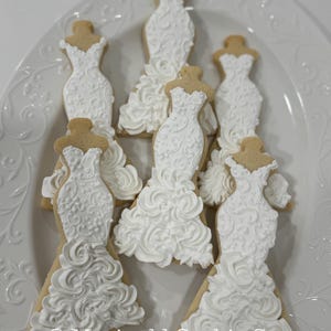 May include: A collection of decorated sugar cookies shaped like wedding dresses. The cookies are iced in white with intricate designs and placed on a white, decorative plate. The cookies have a light brown base.