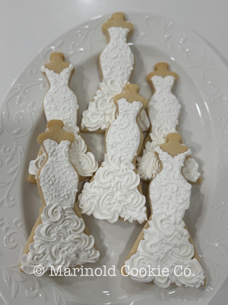 May include: A collection of decorated sugar cookies shaped like wedding dresses. The cookies are iced in white with intricate designs and placed on a white, decorative plate. The cookies have a light brown base.