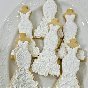May include: A white, iced sugar cookie shaped like a wedding dress, with intricate floral detailing. The cookies are arranged on a white, decorative plate. The text "Marinold Cookie Co." is visible at the bottom of the image. A sweet treat for a bridal shower or wedding.