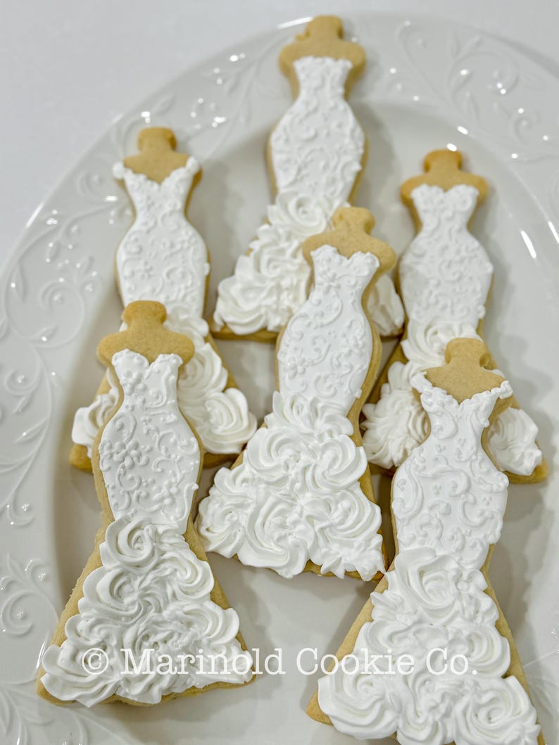 May include: A white, iced sugar cookie shaped like a wedding dress, with intricate floral detailing. The cookies are arranged on a white, decorative plate. The text "Marinold Cookie Co." is visible at the bottom of the image. A sweet treat for a bridal shower or wedding.