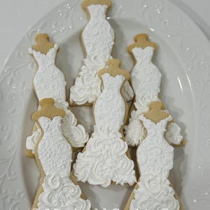 May include: A collection of wedding dress-shaped cookies, decorated with white icing, arranged on a white, decorative plate. The cookies feature intricate details and a textured appearance. The text "Marinold Cookie Co." is visible.