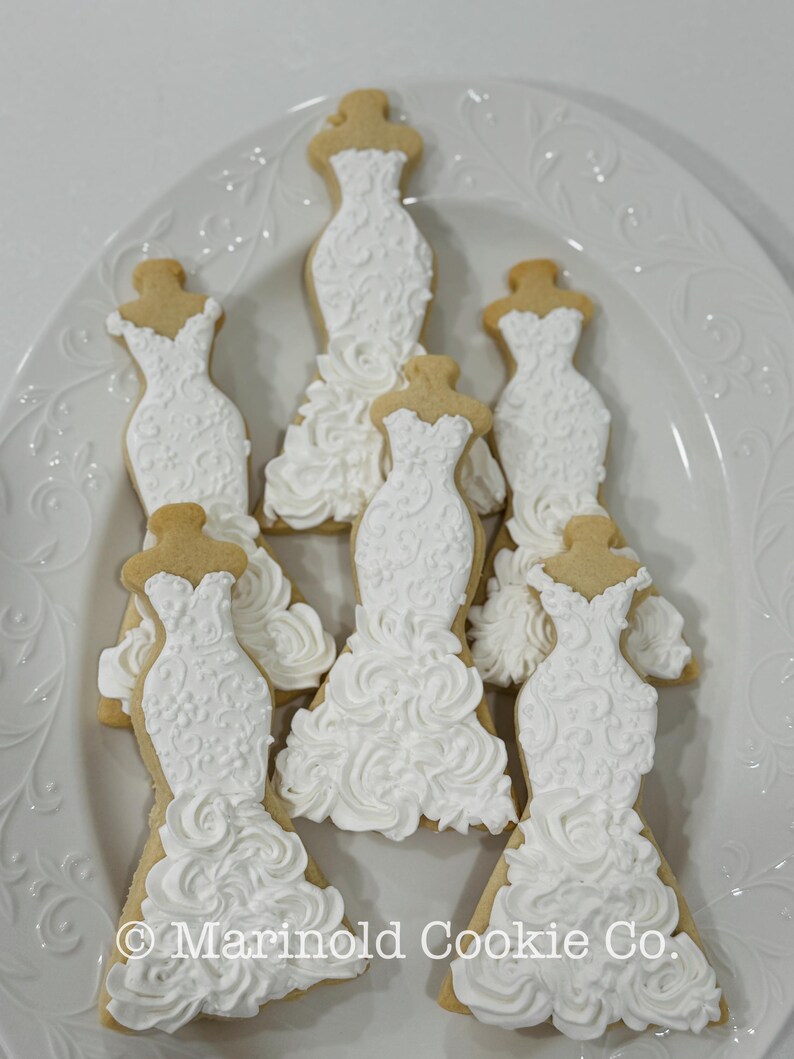 May include: A collection of wedding dress-shaped cookies, decorated with white icing, arranged on a white, decorative plate. The cookies feature intricate details and a textured appearance. The text "Marinold Cookie Co." is visible.
