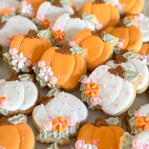 May include: Close-up of pumpkin-shaped cookies, some orange and some white, decorated with floral icing. The cookies have brown stems and are arranged on a light-colored surface. The text "Marinold Cookie Co." is visible.