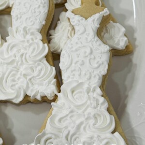 May include: Close-up of several wedding dress-shaped cookies. The cookies are decorated with white frosting, featuring intricate details and a textured, ruffled skirt. The cookies are on a white plate. The text "Marinold Cookie Co." is visible.