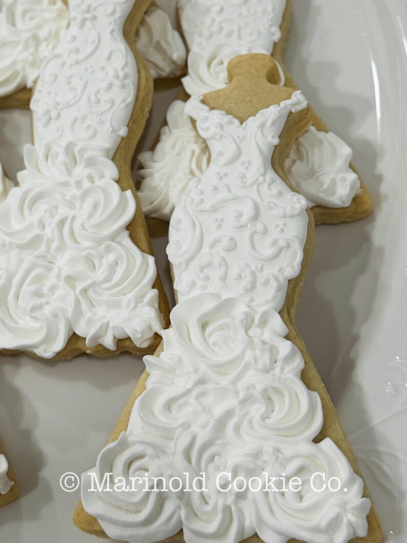 May include: Close-up of several wedding dress-shaped cookies. The cookies are decorated with white frosting, featuring intricate details and a textured, ruffled skirt. The cookies are on a white plate. The text "Marinold Cookie Co." is visible.