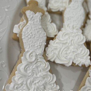 May include: Close-up of several wedding dress-shaped cookies. The cookies are decorated with white icing, featuring intricate details and a ruffled skirt design. The cookies are on a white plate. The text "Marinold Cookie Co." is visible.