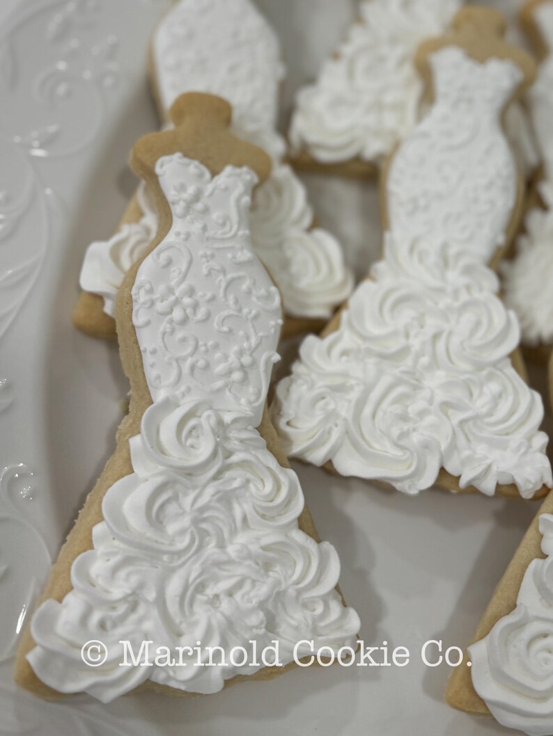 May include: Close-up of several wedding dress-shaped cookies. The cookies are decorated with white icing, featuring intricate details and a ruffled skirt design. The cookies are on a white plate. The text "Marinold Cookie Co." is visible.