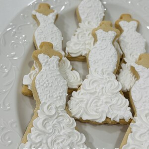 May include: A white plate holds several sugar cookies shaped like wedding dresses. The cookies have a light brown base and are decorated with white icing, featuring intricate details and a ruffled skirt design. The text "Marinold Cookie Co." is visible.
