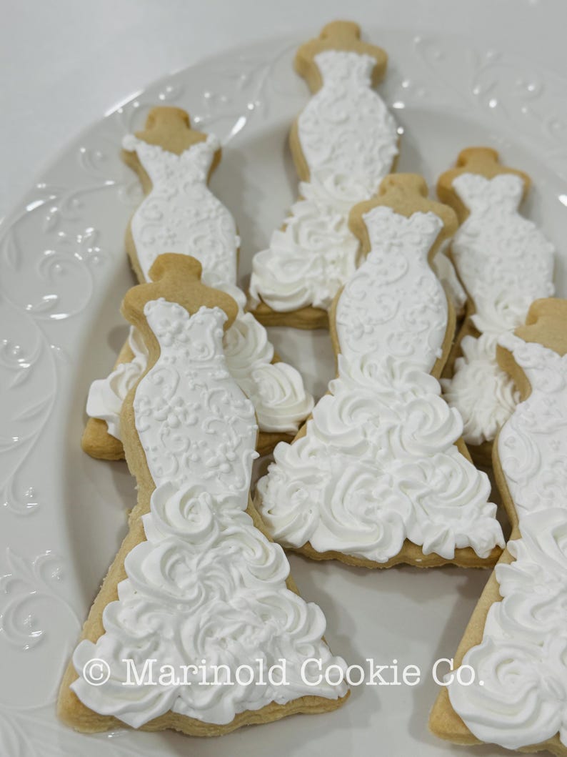 May include: A white plate holds several sugar cookies shaped like wedding dresses. The cookies have a light brown base and are decorated with white icing, featuring intricate details and a ruffled skirt design. The text "Marinold Cookie Co." is visible.
