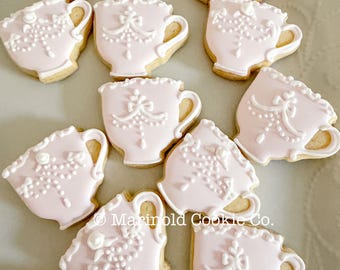 May include: A collection of pink teacup-shaped cookies with white decorative icing. The cookies have a light brown base and are arranged on a light-colored surface. The cookies are decorated with floral and beaded designs.