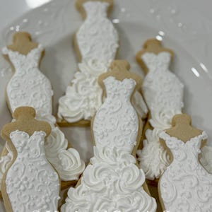 May include: A collection of decorated sugar cookies shaped like wedding dresses. The cookies have a light brown base with white icing details, including floral patterns and ruffled skirts. The cookies are arranged on a white plate.