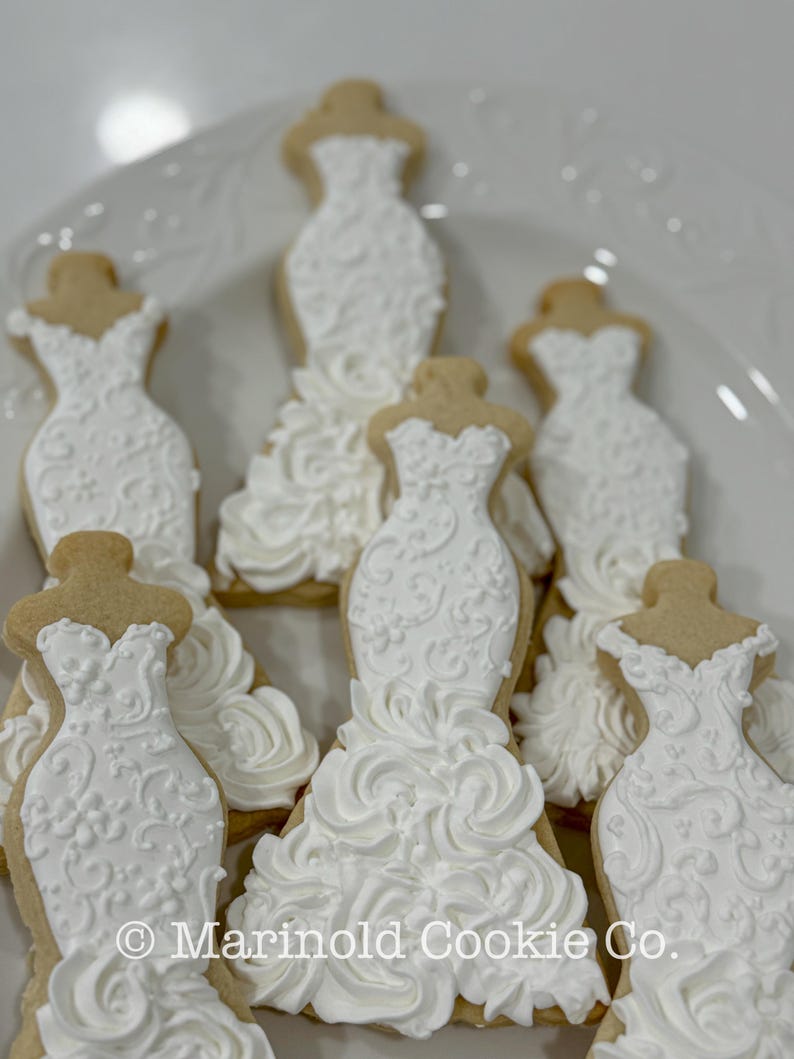 May include: A collection of decorated sugar cookies shaped like wedding dresses. The cookies have a light brown base with white icing details, including floral patterns and ruffled skirts. The cookies are arranged on a white plate.