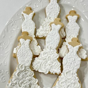 May include: A white platter holds several decorated sugar cookies shaped like wedding dresses. The dresses are iced with white frosting, featuring intricate details and textures. The cookies have a light brown base.