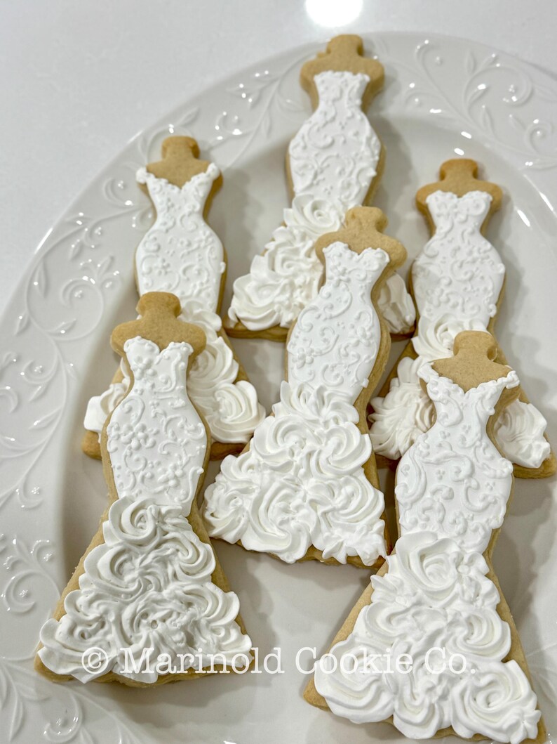 May include: A white platter holds several decorated sugar cookies shaped like wedding dresses. The dresses are iced with white frosting, featuring intricate details and textures. The cookies have a light brown base.