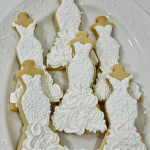 May include: A collection of wedding dress-shaped cookies, iced in white with intricate details, arranged on a white, decorative plate. The cookies have a light brown base and are decorated with white icing, resembling bridal gowns. The text "Marinold Cookie Co." is visible.
