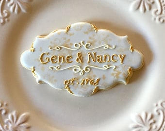 May include: A decorated cookie with the names "Gene & Nancy" and the date "est. 1968" in gold lettering. The cookie has a white background with a gold floral pattern and is edged in gold. It rests on a white, ornate plate.