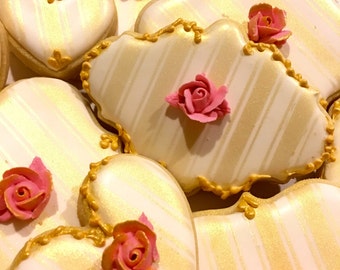 May include: Close-up of decorated cookies in various shapes, including hearts and cloud-like forms. The cookies are white with gold stripes and gold trim, and topped with pink and orange sugar roses. The cookies are arranged on a white plate.