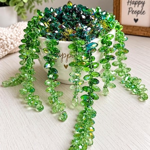 May include: A decorative plant sculpture made of faceted green crystal beads arranged in long, trailing strands. The beads are housed in a white ceramic bowl with the word "happy" and a small heart printed on the front in gold lettering.