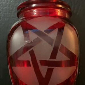 Peut inclure: Un pot en verre rouge avec un motif de pentagramme givré. Le pot a un couvercle de forme hexagonale.