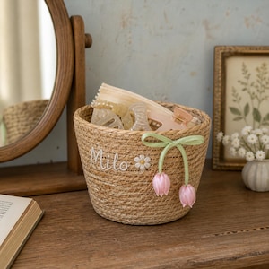 May include: A round, woven tan storage basket featuring the embroidered name Milo, a small white daisy appliqué, and a light green ribbon tied in a bow with two dangling pink tulip charms. The basket contains several neutral-toned hair clips.