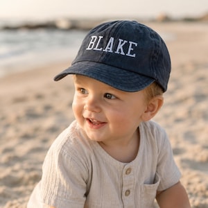 Könnte beinhalten: Eine marineblaue Baseballkappe mit dem Namen BLAKE, der in weißen Großbuchstaben auf die Vorderseite gestickt ist. Die Kappe hat einen gebogenen Schirm und eine strukturierte Krone aus einfarbigem Stoff mit sichtbaren Nähten.
