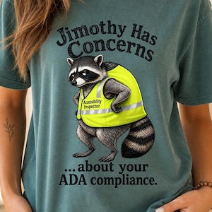 May include: A teal short-sleeve t-shirt featuring a graphic of a raccoon wearing a yellow high-visibility vest labeled Accessibility Inspector. The text reads Jimothy Has Concerns about your ADA compliance in black lettering above and below the raccoon.