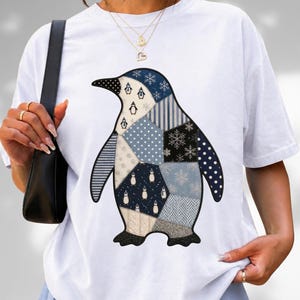 Peut inclure: Un t-shirt blanc à manches courtes arborant un grand motif central représentant un manchot composé de divers motifs en patchwork bleu, blanc et noir, incluant des flocons de neige, des pois, des rayures et de petites silhouettes de manchots.