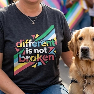 May include: A dark gray short-sleeve t-shirt featuring the text "different is not broken" in colorful letters, set against a background of rainbow-colored geometric shapes. The shirt is worn by a person standing next to a golden retriever dog.
