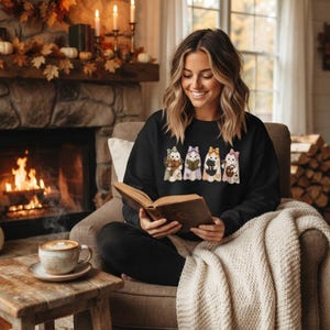 Peut inclure: Un sweat-shirt noir à manches longues orné d'un imprimé graphique représentant quatre fantômes portant des nœuds, tenant chacun des objets différents dont un bouquet, un livre, un chat noir et un panier, tout en lisant un livre ouvert.