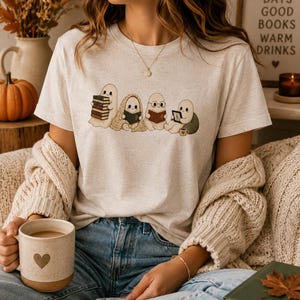 Könnte beinhalten: Ein meliertes, hellbeiges T-Shirt mit einer Grafik von vier kleinen, cartoonartigen Geistern beim Lesen. Ein Geist hält einen Stapel Bücher, ein anderer ist in eine Decke gewickelt, einer trägt eine Brille und der letzte hält ein Tablet.