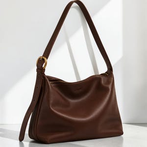 Puede incluir: Un bolso de hombro de color marrón oscuro con acabado liso, que cuenta con una sola correa ajustable sujeta con una hebilla circular de tono latón antiguo. El bolso tiene una silueta superior curva con cierre de cremallera y base estructurada.