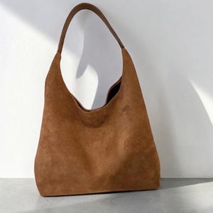 May include: A medium brown hobo bag crafted from a soft, textured suede material. The bag features a single integrated shoulder strap that curves into the main body, creating a slouchy, unstructured silhouette with a wide, open top opening.