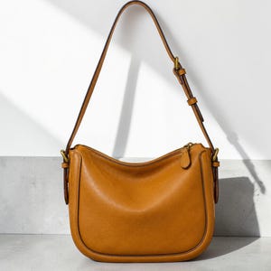 May include: A tan leather shoulder bag with a curved bottom silhouette, featuring a single adjustable strap with a brass buckle, a top zip closure with a matching leather pull tab, and reinforced side panels with brass hardware attachments.