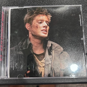 May include: A clear plastic CD jewel case featuring a printed insert of a person with short brown hair, a dirt-smudged face, and a dark jacket. The text "DEAN WINCHESTER" is printed vertically in red along the left side of the case cover.