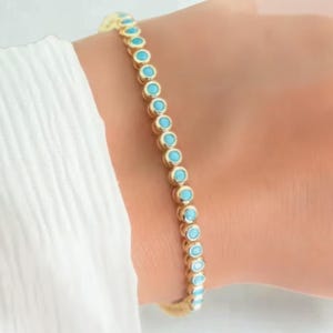 May include: A gold-toned tennis bracelet featuring a continuous row of small, round, bright turquoise-colored stones set in individual metal bezels. The bracelet is worn around the wrist, showcasing a uniform design of alternating gold and blue.