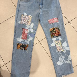 May include: Light blue denim jeans featuring a button fly, belt loops, and five pockets. The legs are decorated with white painted floral motifs and several rectangular fabric patches with various colourful patterns and frayed edges.
