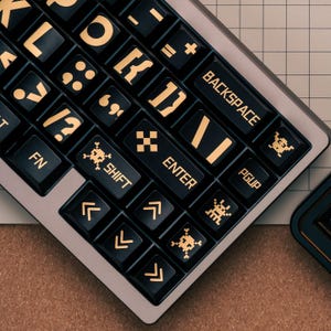 May include: A mechanical keyboard with black keycaps featuring gold-colored legends and pixelated graphics. The keys include text such as BACKSPACE, ENTER, SHIFT, FN, and PGUP, alongside symbols like skulls and arrows, set against a metallic frame.