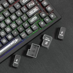 May include: A black mechanical keyboard with backlit keys featuring custom white symbols and text, including ENTER and SHIFT. Several loose, dark gray replacement keycaps are displayed nearby, showing their underside with cross-shaped mounts.