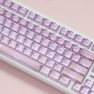 May include: A white mechanical keyboard featuring translucent light pink keycaps with silver metallic edges. The keys display standard alphanumeric characters and function labels. The right shift key includes the text A crash report has been generated.