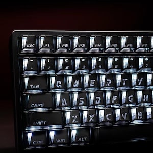 May include: A black mechanical keyboard with translucent keycap sides and white backlit lettering. The keys feature a QWERTY layout with visible labels including ESC, TAB, CAPS, SHIFT, CTRL, WIN, and ALT, illuminated by bright white light.