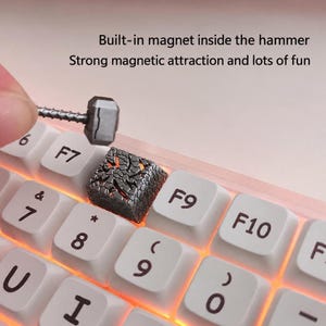 May include: A custom mechanical keyboard keycap featuring a textured, cracked gray surface with orange light glowing from beneath. A small, metallic gray hammer with a built-in magnet is held above the keycap. Text reads: Built-in magnet inside the hammer. Strong magnetic attraction and lots of fun.