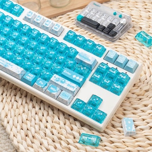 May include: A white mechanical keyboard featuring translucent blue keycaps with white lettering and bow icons. The keyboard includes a separate matching number pad, and several loose keycaps are scattered nearby on the surface.