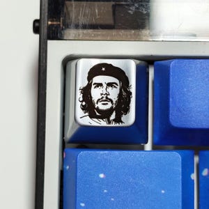 May include: A silver-colored mechanical keyboard keycap featuring a black graphic print of Che Guevara wearing a beret with a star. The keycap is positioned among several solid blue keycaps on a keyboard frame with a black and white border.