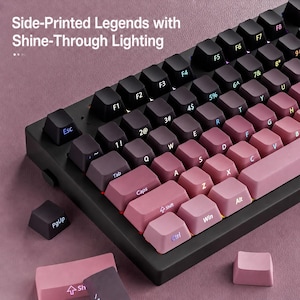 May include: A mechanical keyboard featuring a gradient color scheme transitioning from dark purple to light pink. The keycaps have side-printed legends with shine-through lighting. Additional loose keycaps are visible in the foreground.