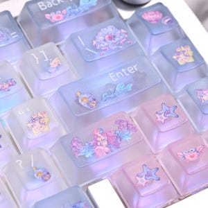 May include: A mechanical keyboard with translucent, frosted keycaps in shades of blue and pink. The keys feature printed illustrations of sea creatures, shells, and bottles. The Enter key is labeled with text, and a silver rotary knob is visible.