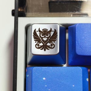 May include: A silver-colored mechanical keyboard keycap featuring a dark brown laser-engraved owl graphic with spread wings. The keycap is positioned among other blue keycaps on a keyboard frame with a black and white border.