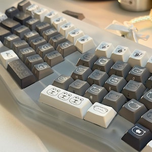 May include: A mechanical keyboard featuring a gray and white color scheme with custom keycaps. The keys are decorated with various line-art illustrations of animal faces, including cats and frogs. The keyboard has a split, ergonomic layout.